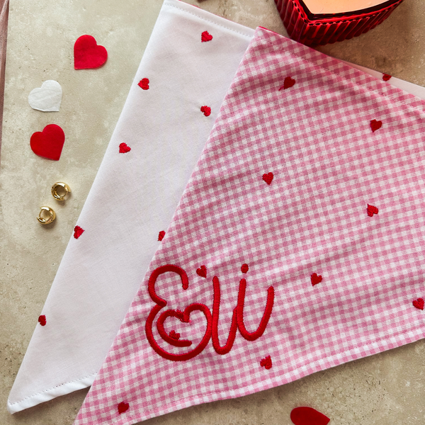 Two fabric triangles, one white with red hearts and one pink gingham with 'Eli' embroidered, on a beige surface.
